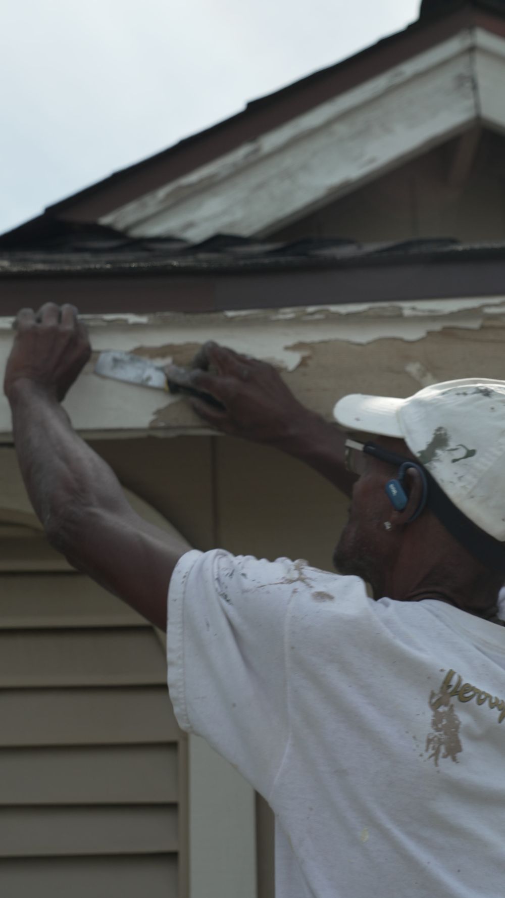 Crew member scraping large flakes of old paint from exterior wood trim beneath the roofline.